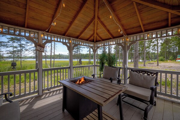 A covered outdoor seating area with a fire pit, wooden furniture, and a view of a lake.