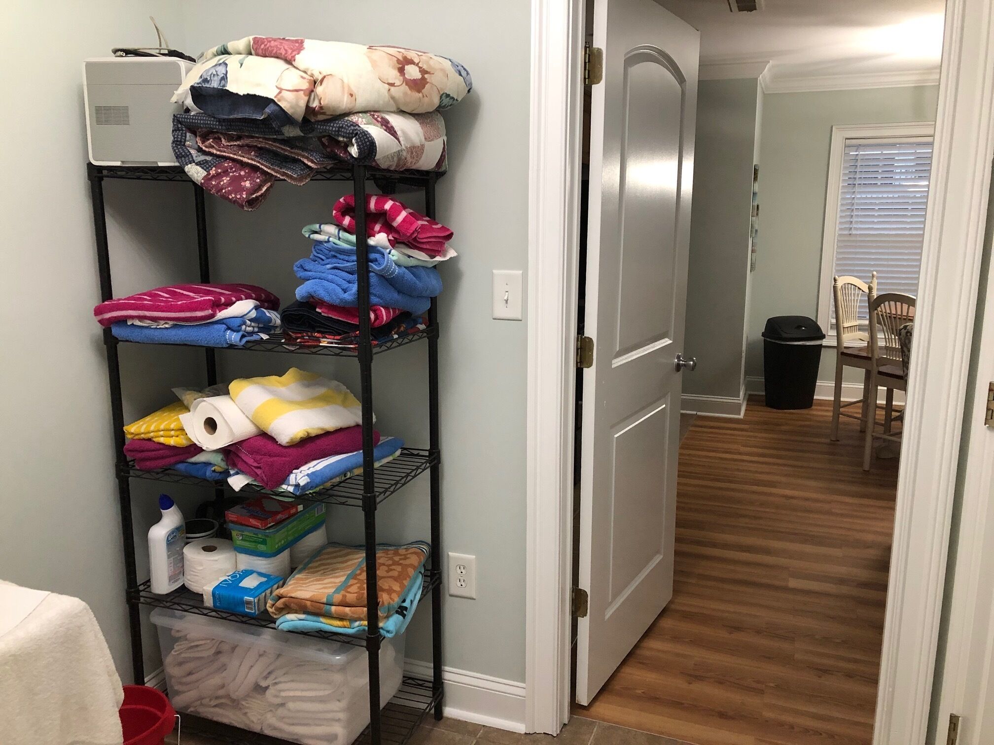 A hallway with a metal shelf unit filled with folded blankets and towels, a trash can, and a closed door.