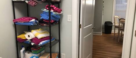 A hallway with a metal shelf unit filled with folded blankets and towels, a trash can, and a closed door.