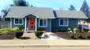 A single-story house with a red door, a small porch, and a well-maintained front yard with plants and mulch.