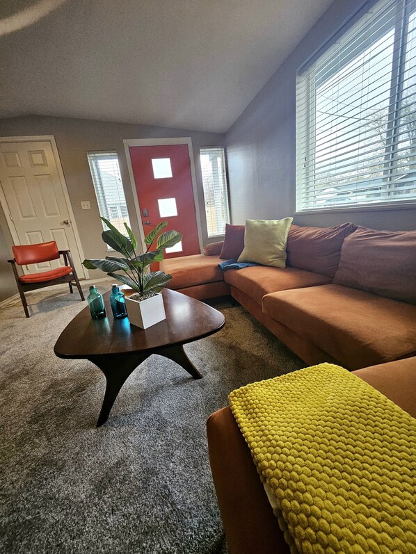 A living room with a brown sofa, a red door, a window with blinds, and a plant on a coffee table.