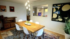 A dining area with a wooden table, white chairs, a hanging light fixture, a mirror, and a large abstract painting.