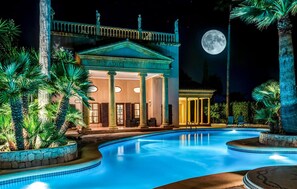 Vista de una piscina con un hotel de fachada clásica, una luna en el cielo y palmeras.