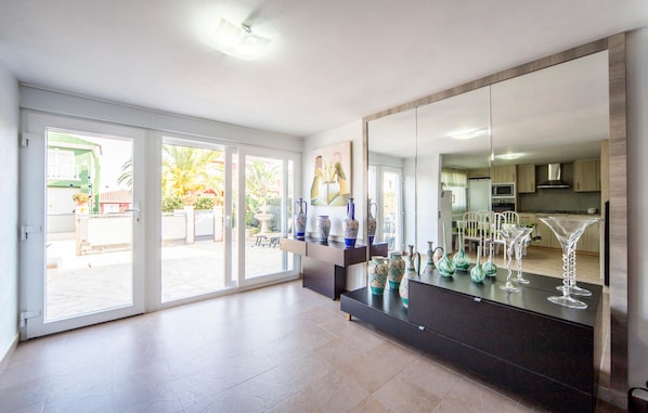 A spacious living area with a large sliding glass door leading to a balcony, a dark wooden cabinet with decorative vases, and a kitchen visible through the glass door. - Villa Carolina Peñíscola (Peñíscola)