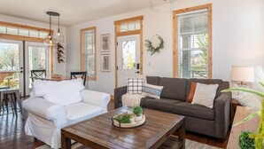 Sala de estar com um sofá branco, um sofá escuro e uma mesa de centro de madeira com uma planta e objetos decorativos. Há vigas de madeira no teto, uma porta com uma guirlanda e uma janela com persianas.