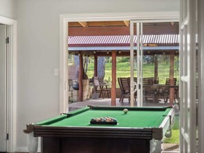 Una mesa de billar en una habitación con un amplio ventanal corredizo que da a un patio exterior con sillas y una mesa.