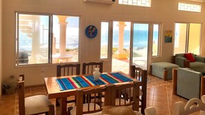 Un vaste espace de vie comprenant une table à manger, des chaises, un ventilateur de plafond et de grandes fenêtres offrant une vue sur l’océan.