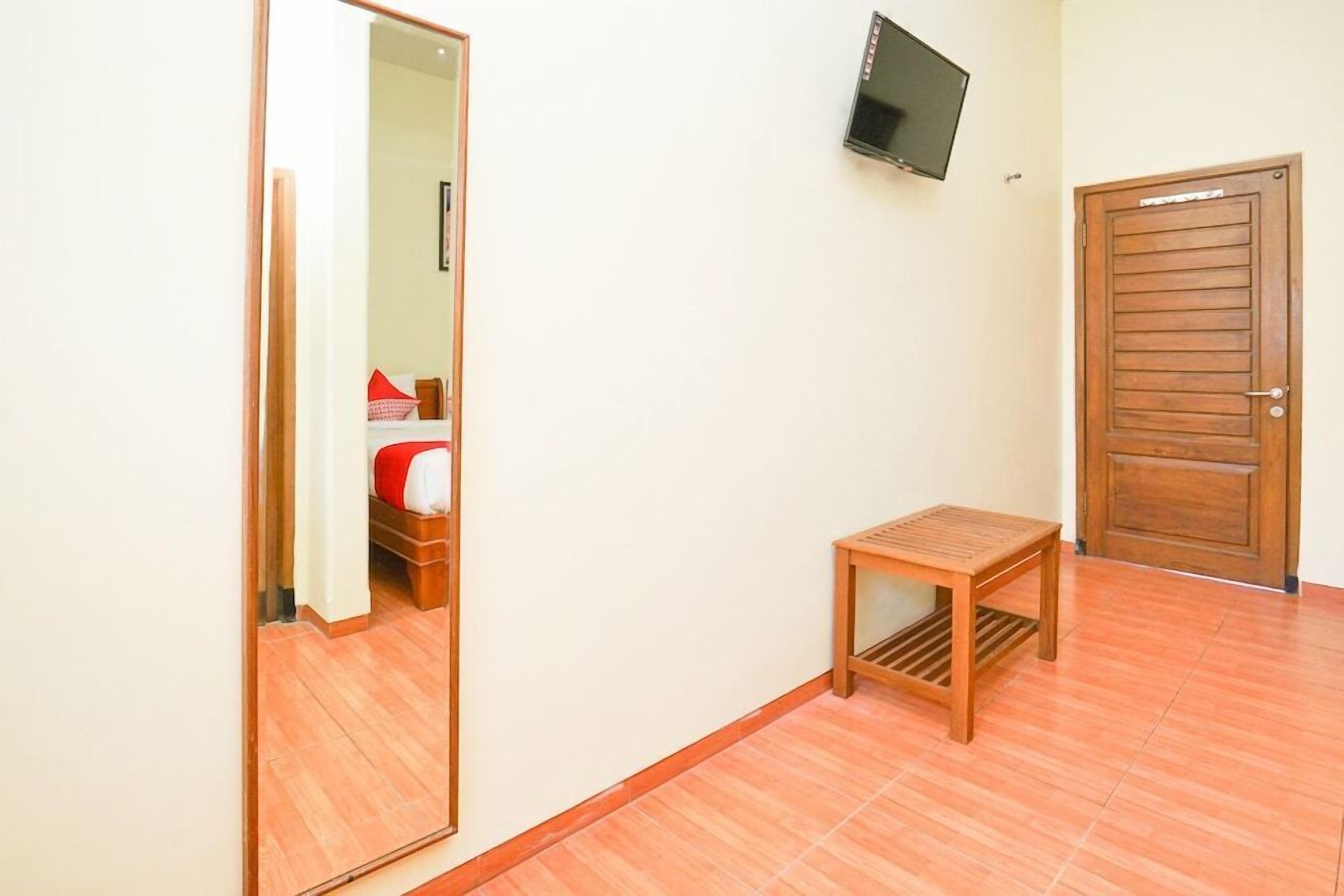 A room with a wooden door, a small wooden table, a television mounted on the wall, and a mirror reflecting part of the room.