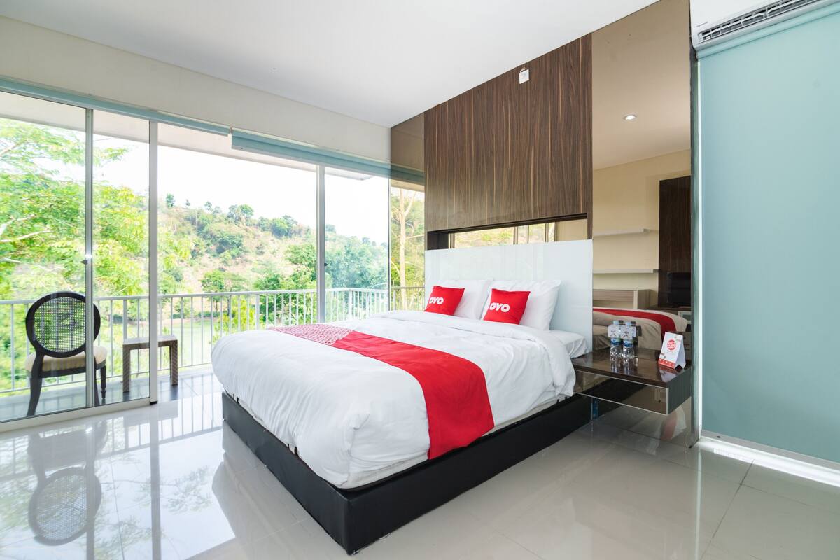 A modern hotel room with a large bed, a balcony with a chair, and a view of greenery.