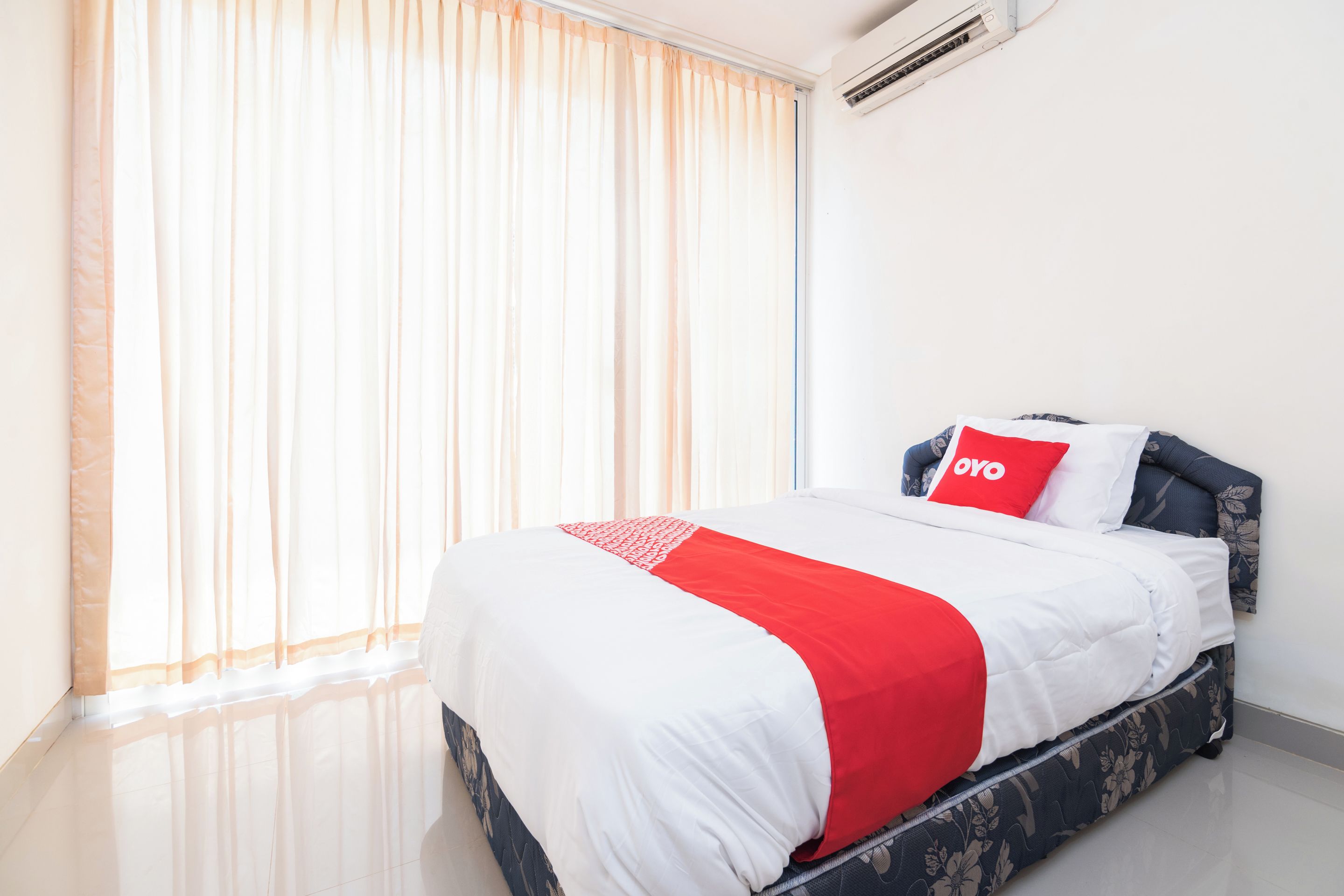 A hotel room with a bed, a window with sheer curtains, an air conditioning unit, and a floral-patterned bed skirt.