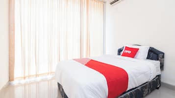 A hotel room with a bed, a window with sheer curtains, an air conditioning unit, and a floral-patterned bed skirt.