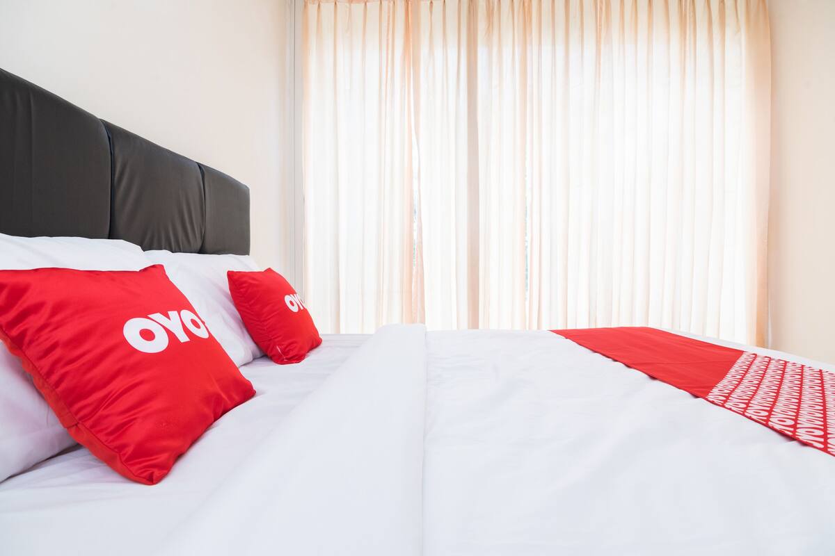 A hotel room with a bed featuring white bedding and red pillows with a logo, a dark headboard, and light curtains.
