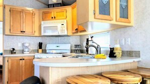 A kitchen with wooden cabinets, a microwave, a stove, and a sink. There are stools and a trash bin.