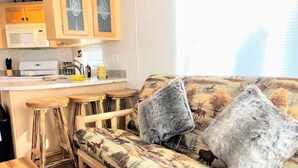 A cozy living area with a sofa, two stools, a wooden table, and a kitchen area with a microwave and cabinets.