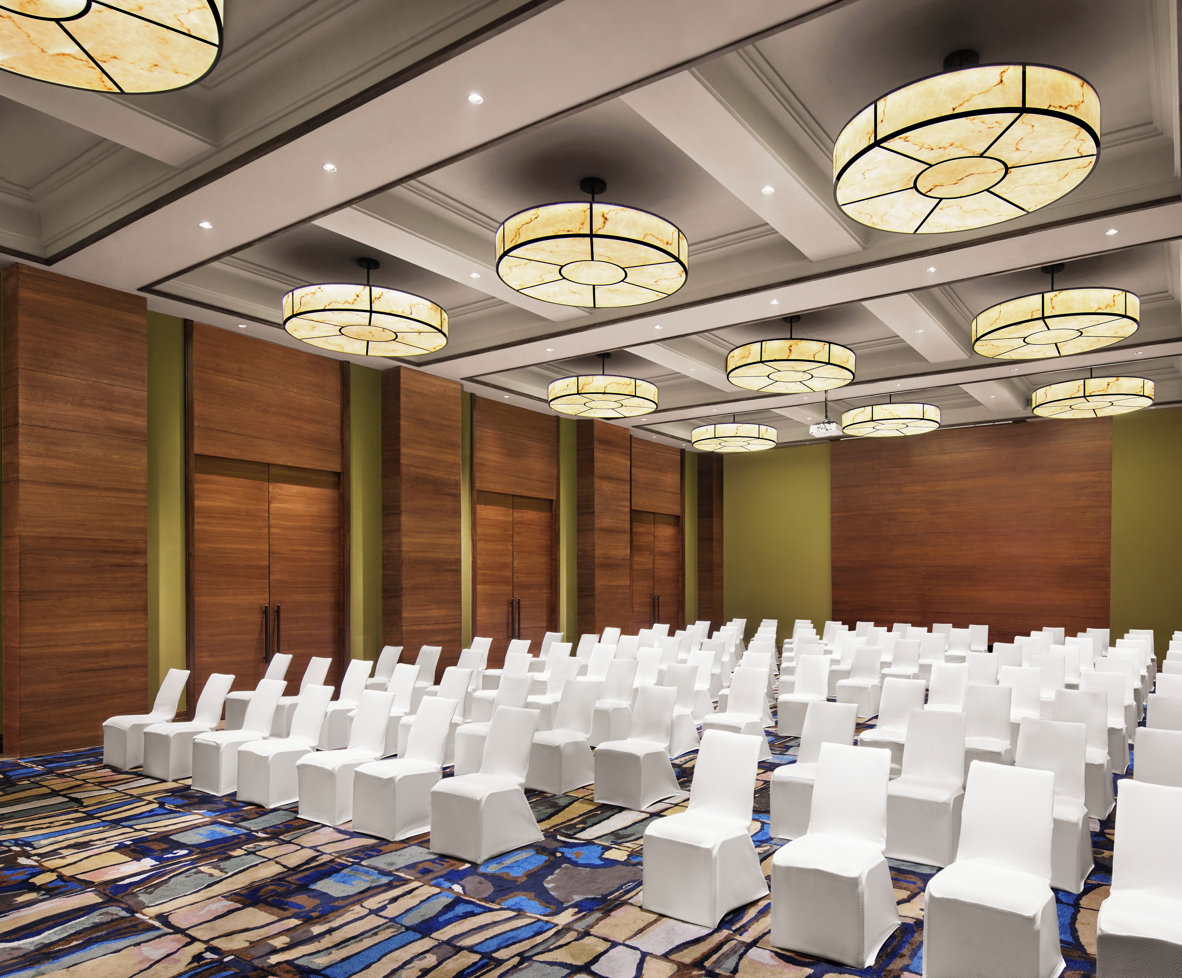 A conference room with rows of white chairs, wooden panel walls, and multiple ceiling chandeliers.