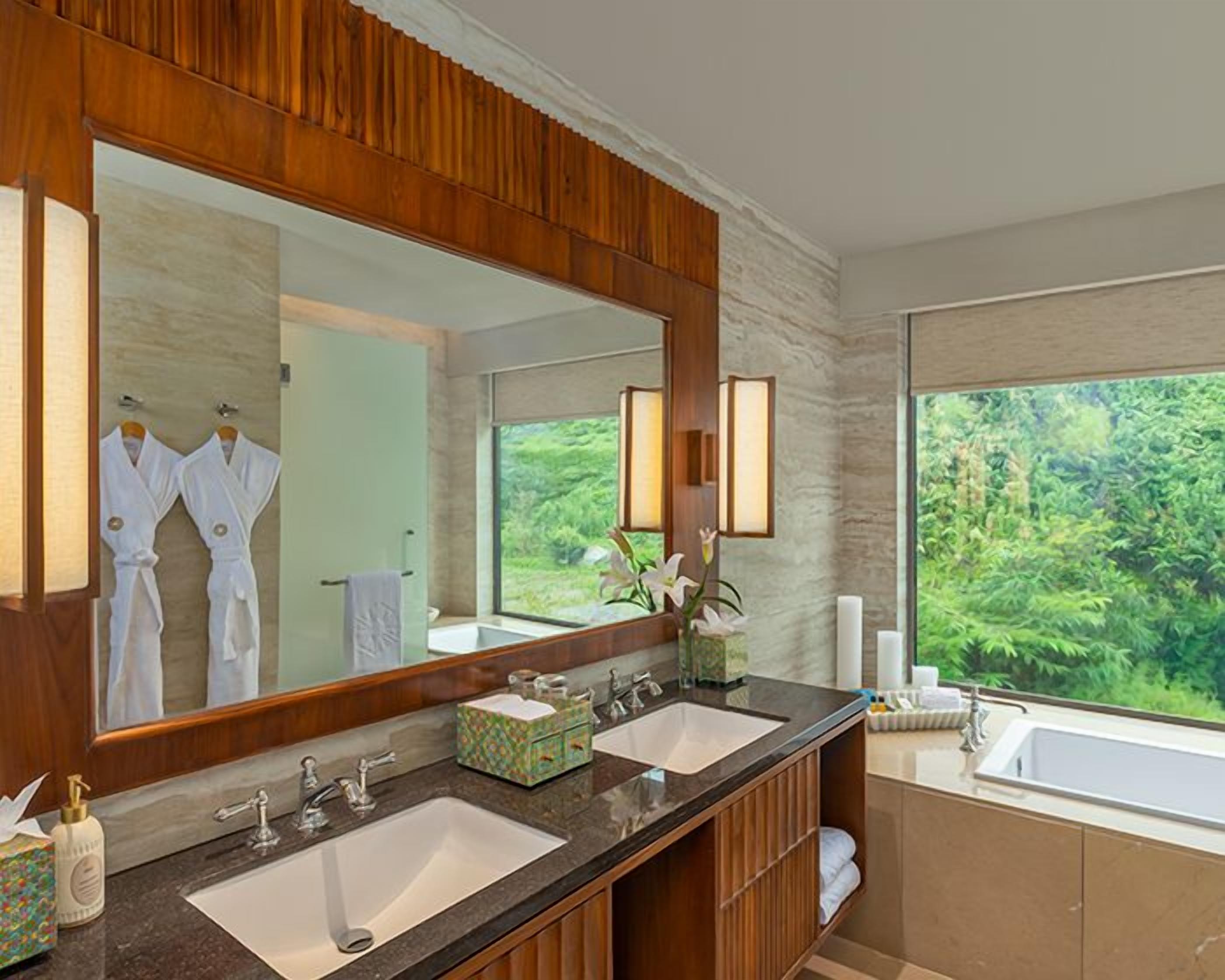 Une salle de bain avec deux lavabos, un grand miroir et une vue sur la verdure à l’extérieur.