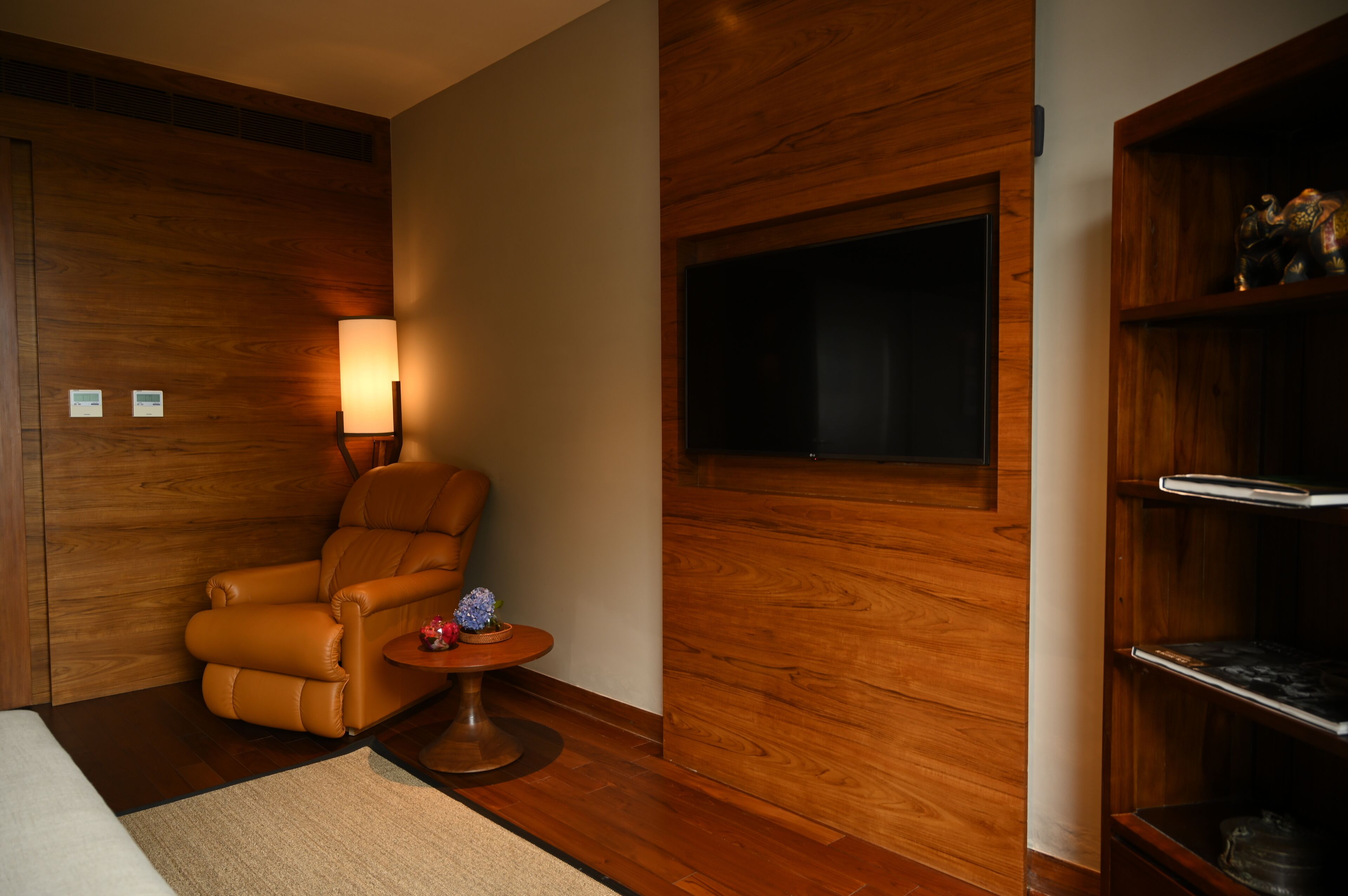Una habitación con pared de madera, un sillón de cuero, una mesita redonda con una lámpara y un televisor montado en la pared.