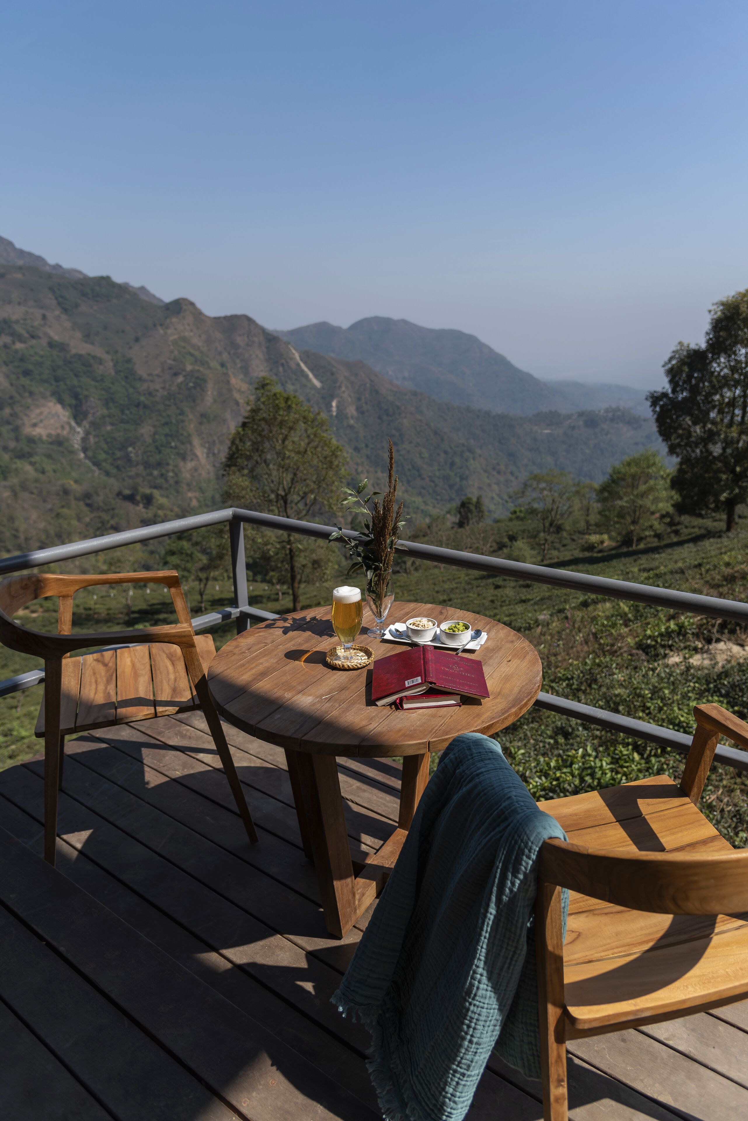 A wooden deck with a round table set for two, a book, and a glass of beer.