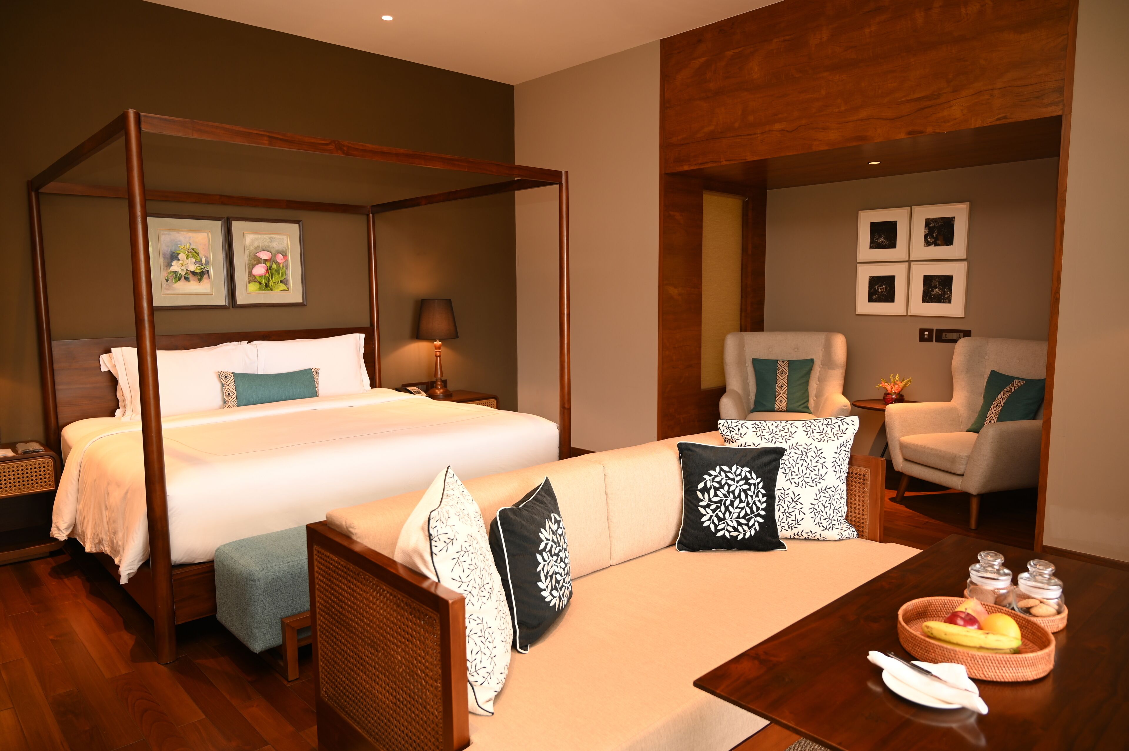 A four-poster bed with a canopy, a sofa with patterned cushions, a small table with fruit and condiments, and a chair with a cushion.