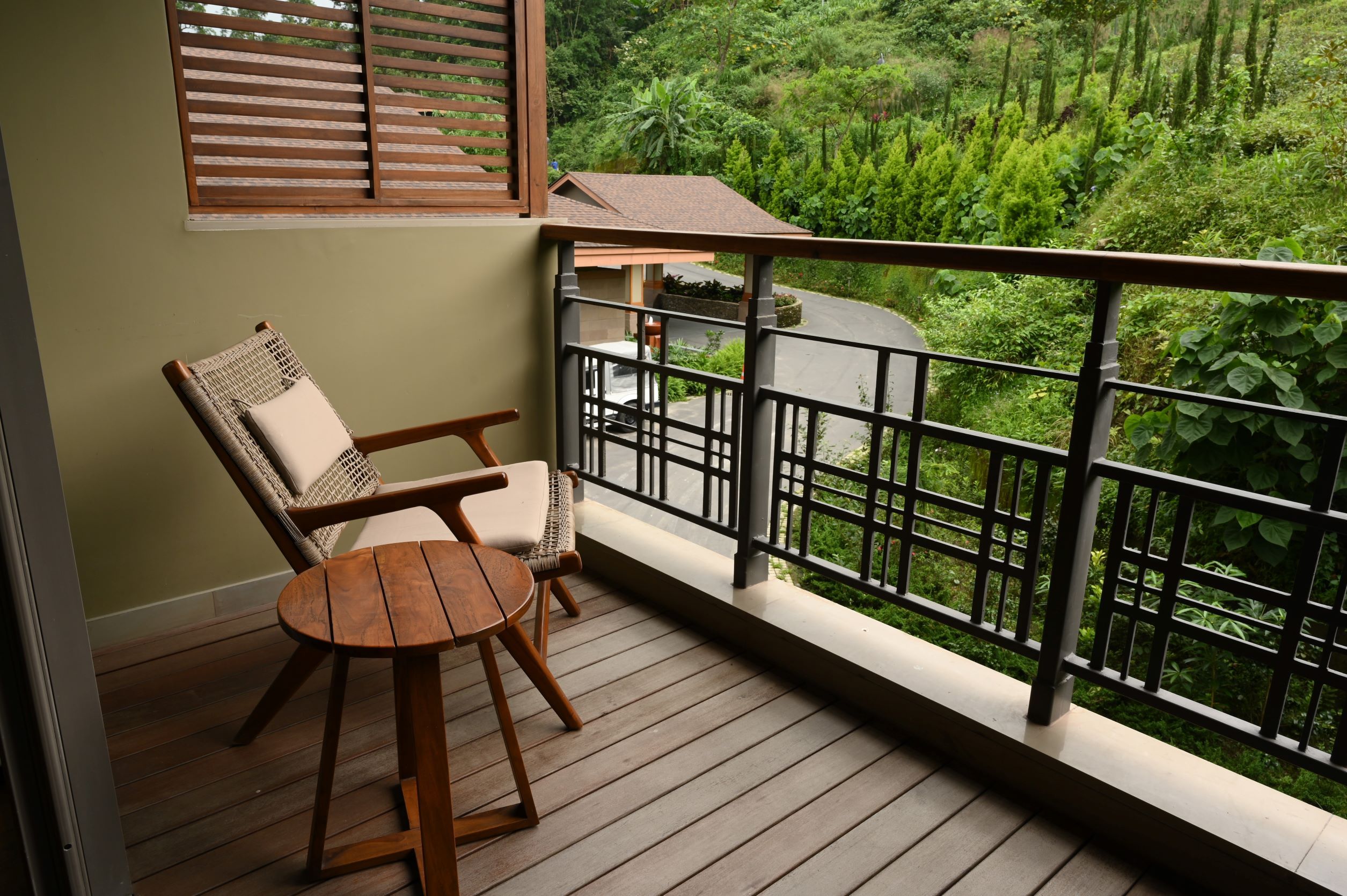 Un balcón con mobiliario de madera, vista a un paisaje verde exuberante y un edificio al fondo.