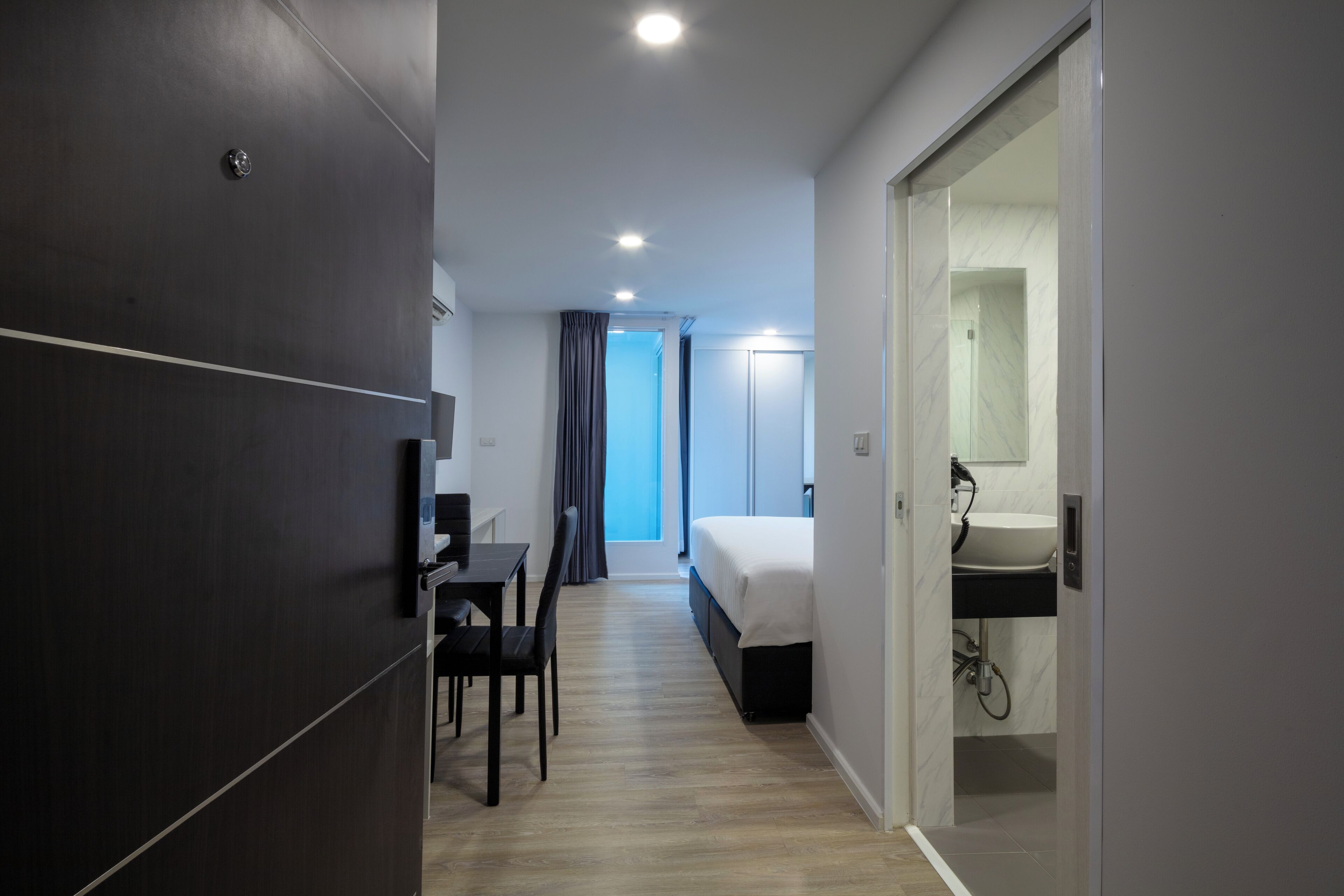Une chambre d’hôtel moderne, équipée d’un lit, d’un bureau et d’une chaise. La salle de bain comprend un lavabo et un miroir. La chambre a un plancher en bois et offre une vue sur l’extérieur.