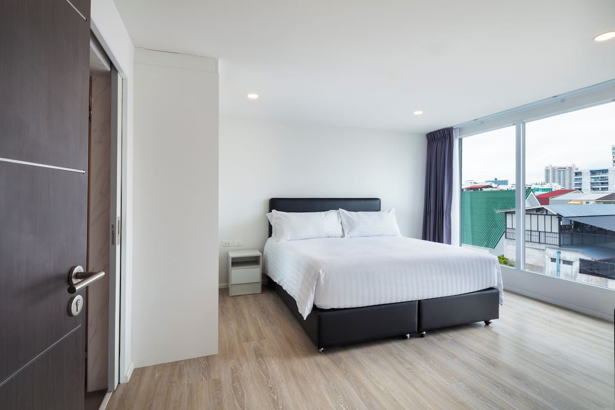 A modern hotel room with a large bed, a nightstand, a wardrobe, and a view of the city through a large window.