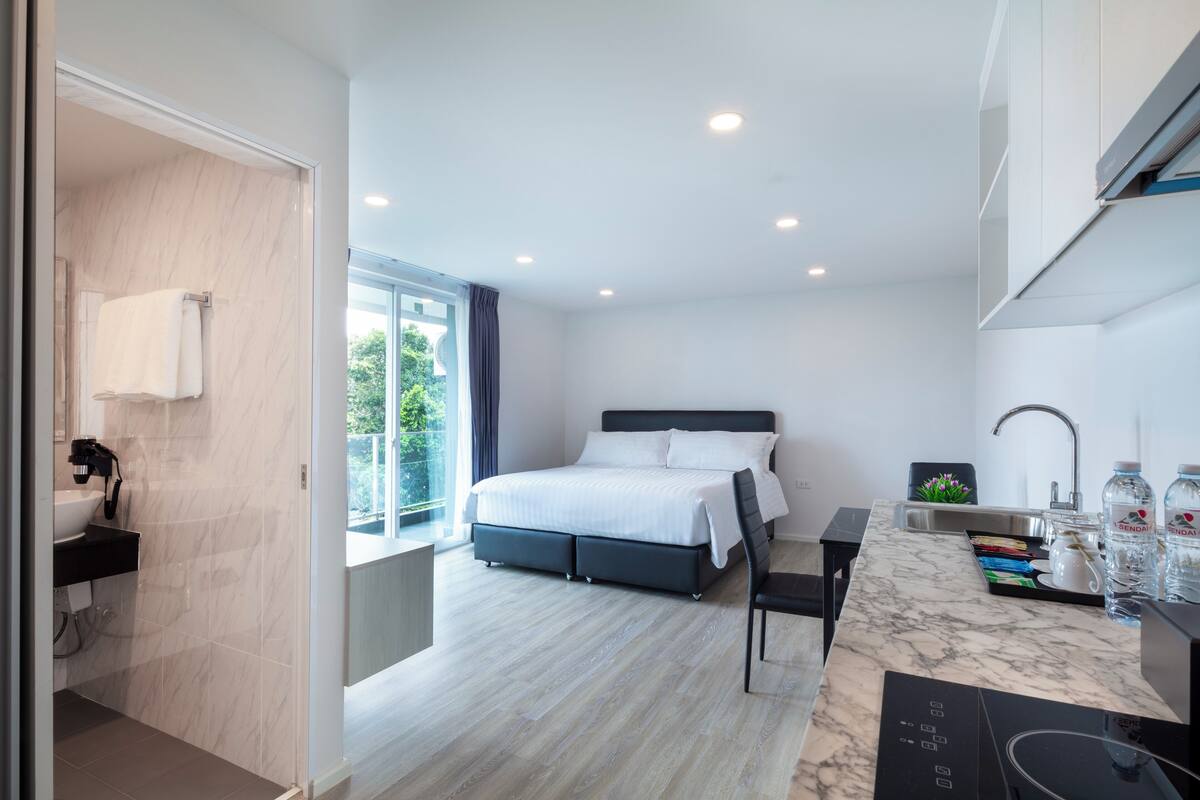 A modern bedroom with a large bed, a bathroom with a sink, and a view of greenery outside.