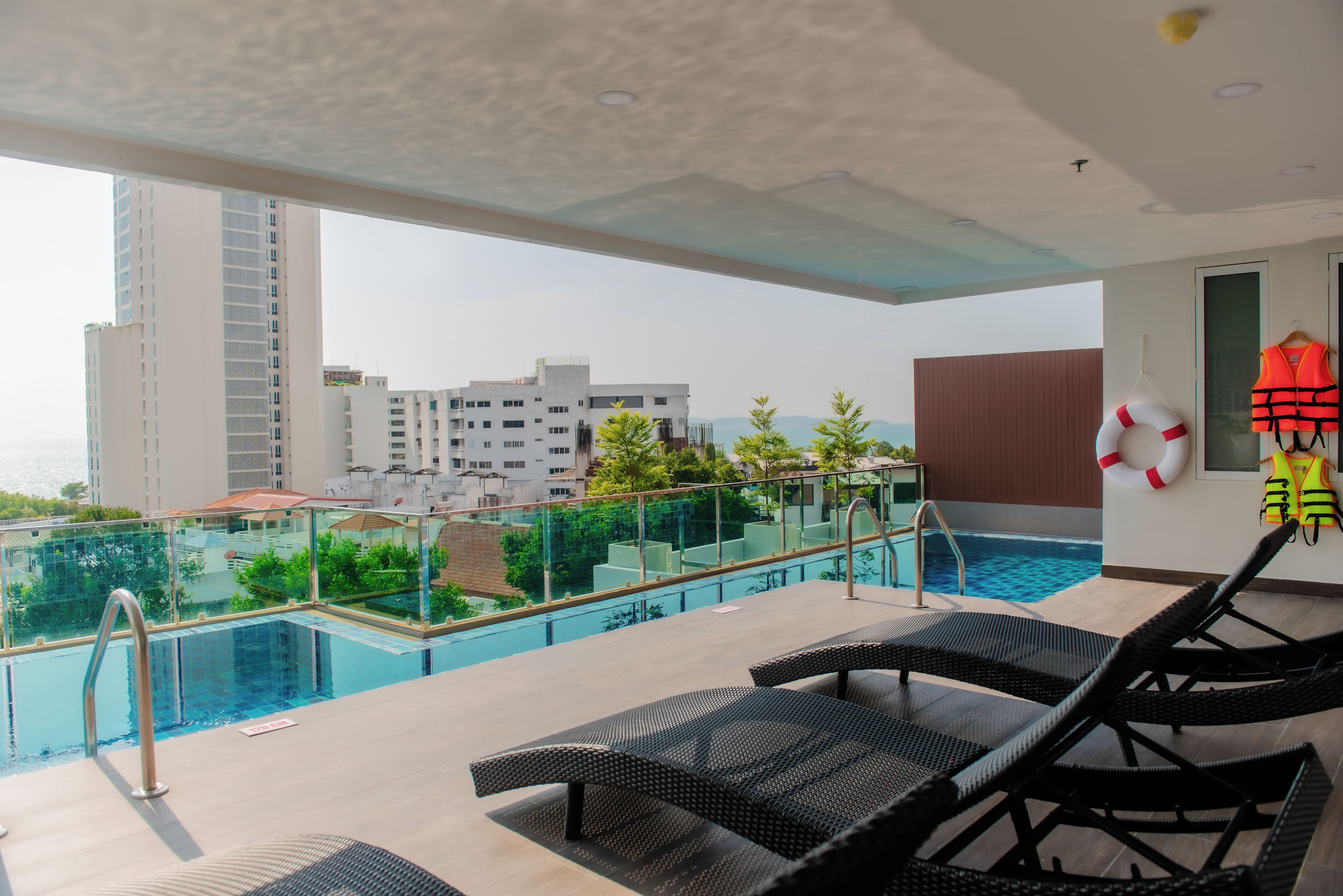 A rooftop pool area with lounge chairs, a lifebuoy, and a view of buildings and greenery.