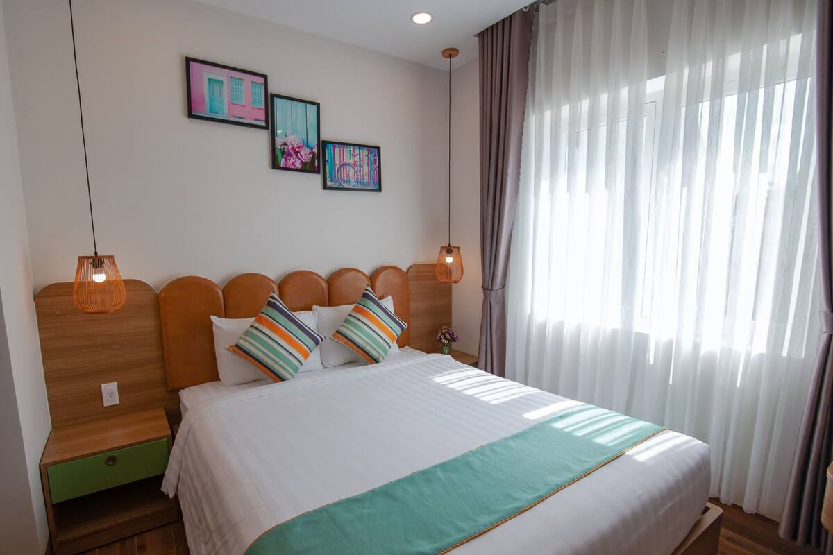 A bedroom with a bed, wooden headboard, two bedside tables, a nightstand, a green throw blanket, and a window with curtains.