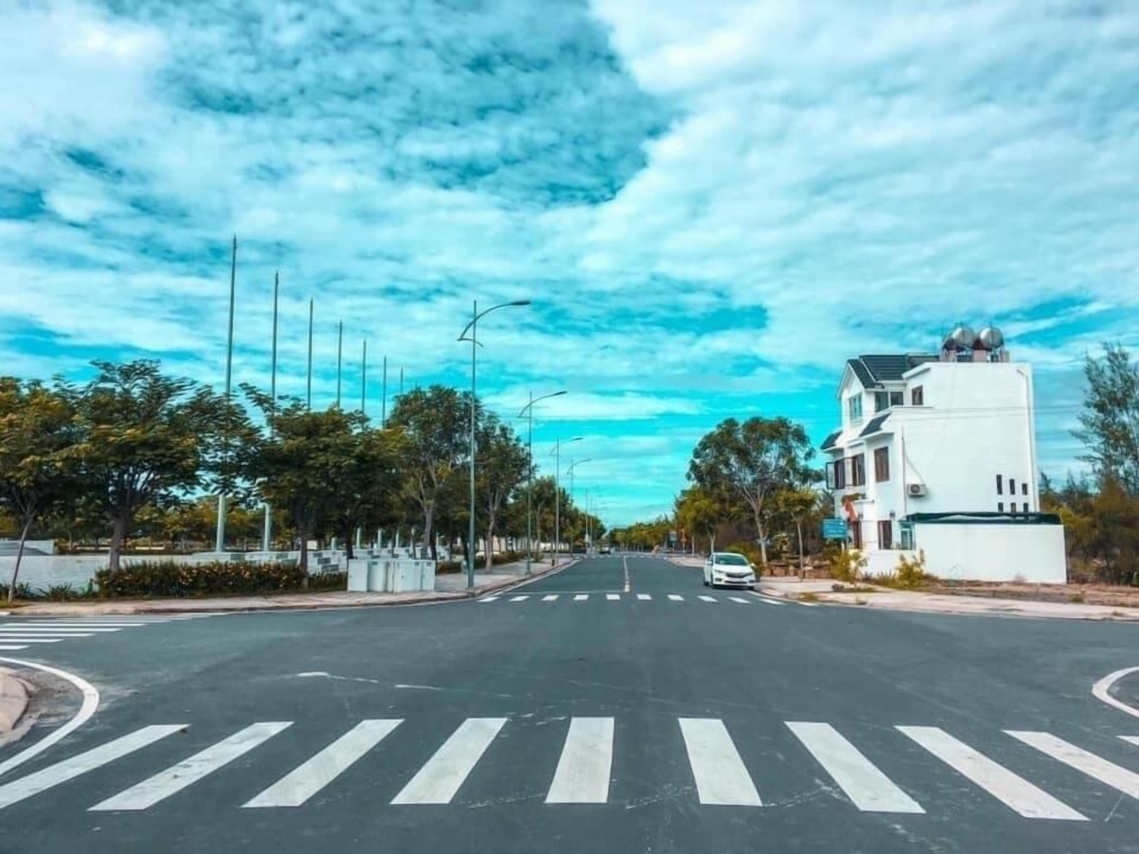 A road with a pedestrian crossing, trees, and a white building with a unique architectural design.