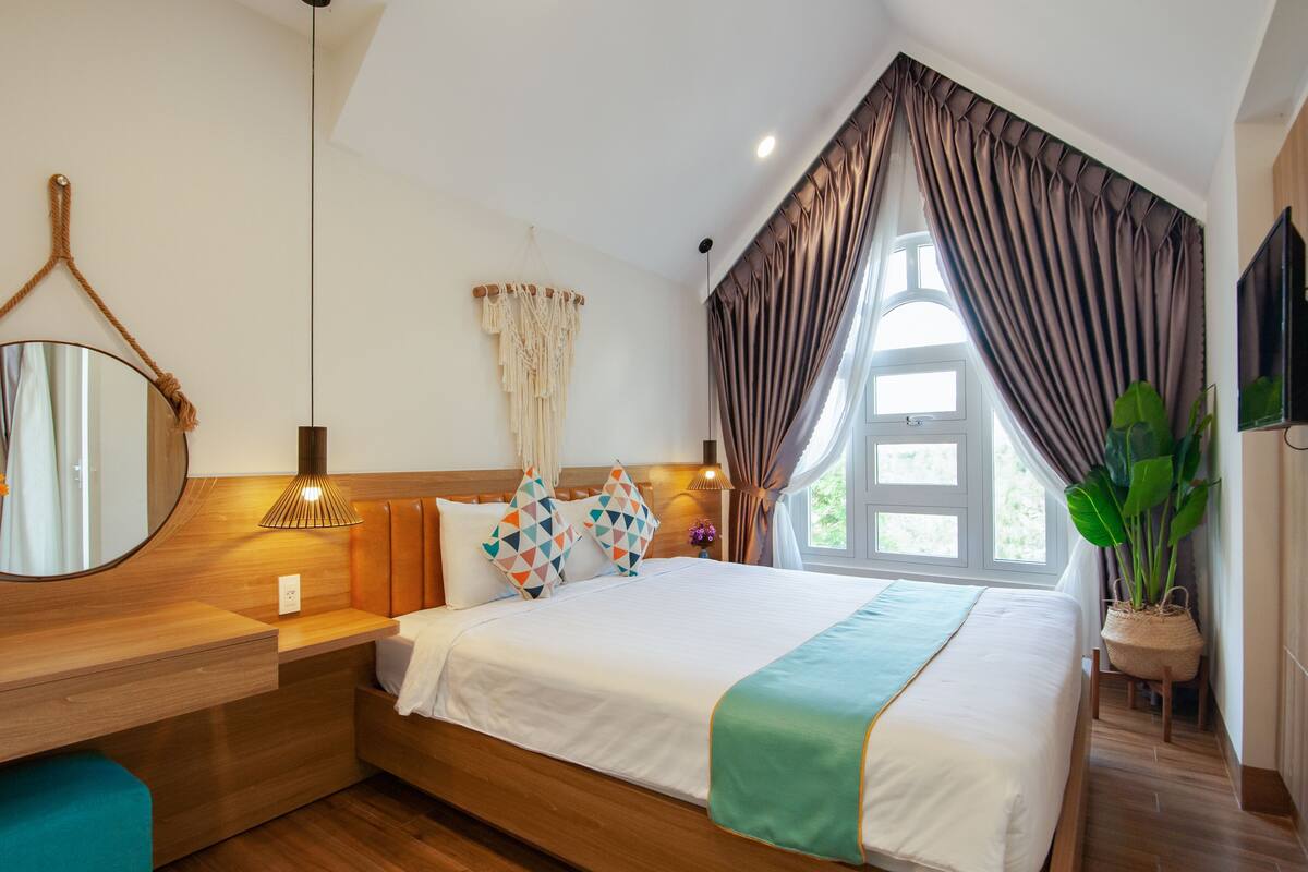 A modern bedroom with a wooden headboard, a round mirror, a bedside table, a bed with a white and green bedspread, and a large window with curtains.