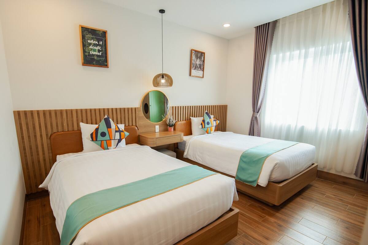 A hotel room with two beds, a wooden headboard, a bedside table with a mirror, a small plant, and a framed picture on the wall.