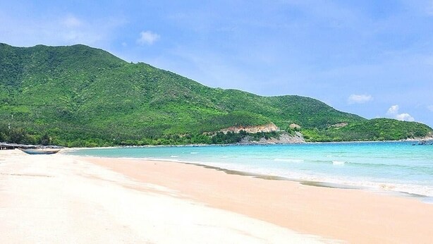 A beach with clear turquoise water, a sandy shore, and a green hill in the background. -  The View Hotel (Cam Lam)