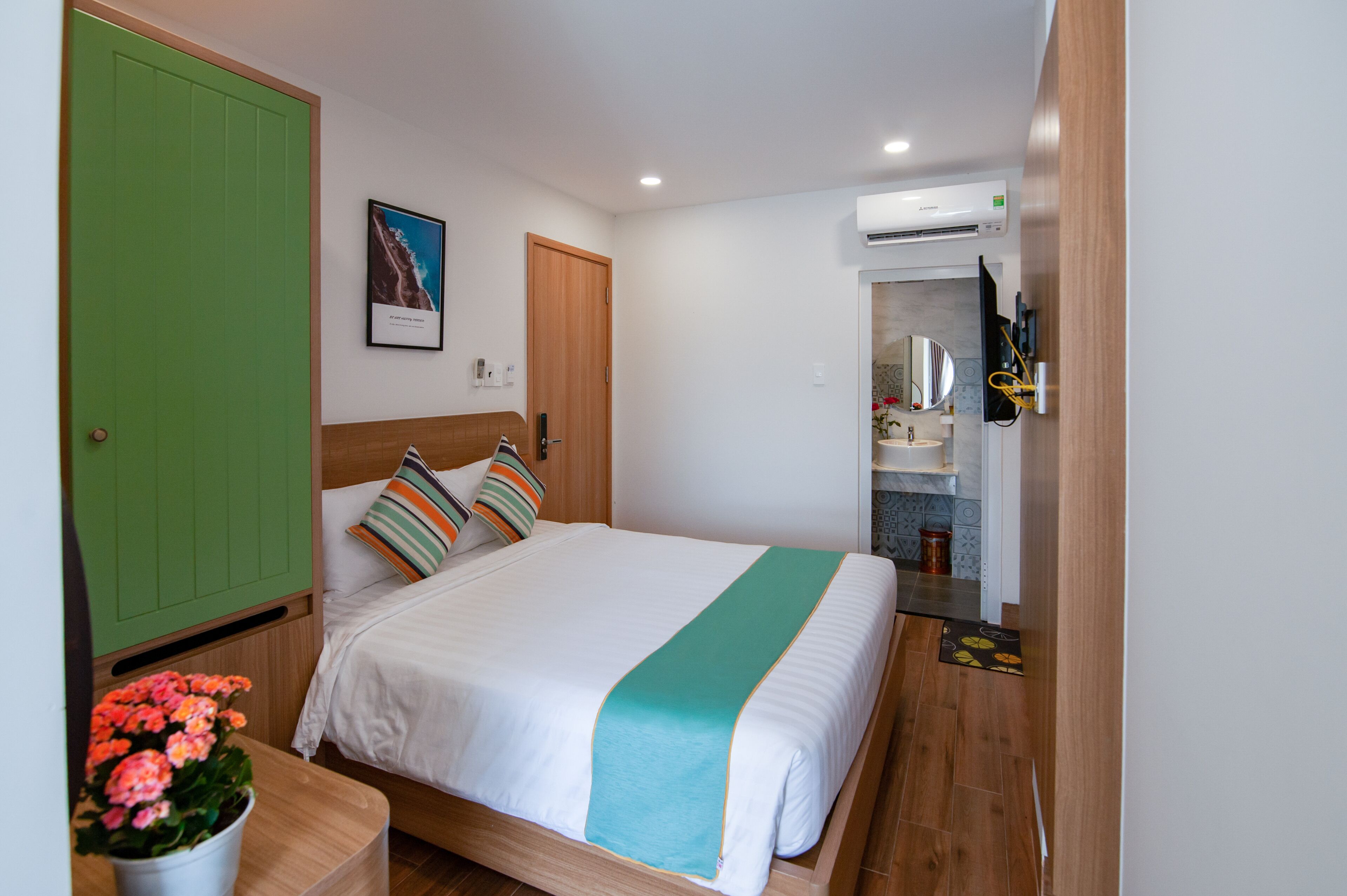 Habitación de hotel con cama, armario verde, baño con lavabo y espejo, y una planta en maceta.