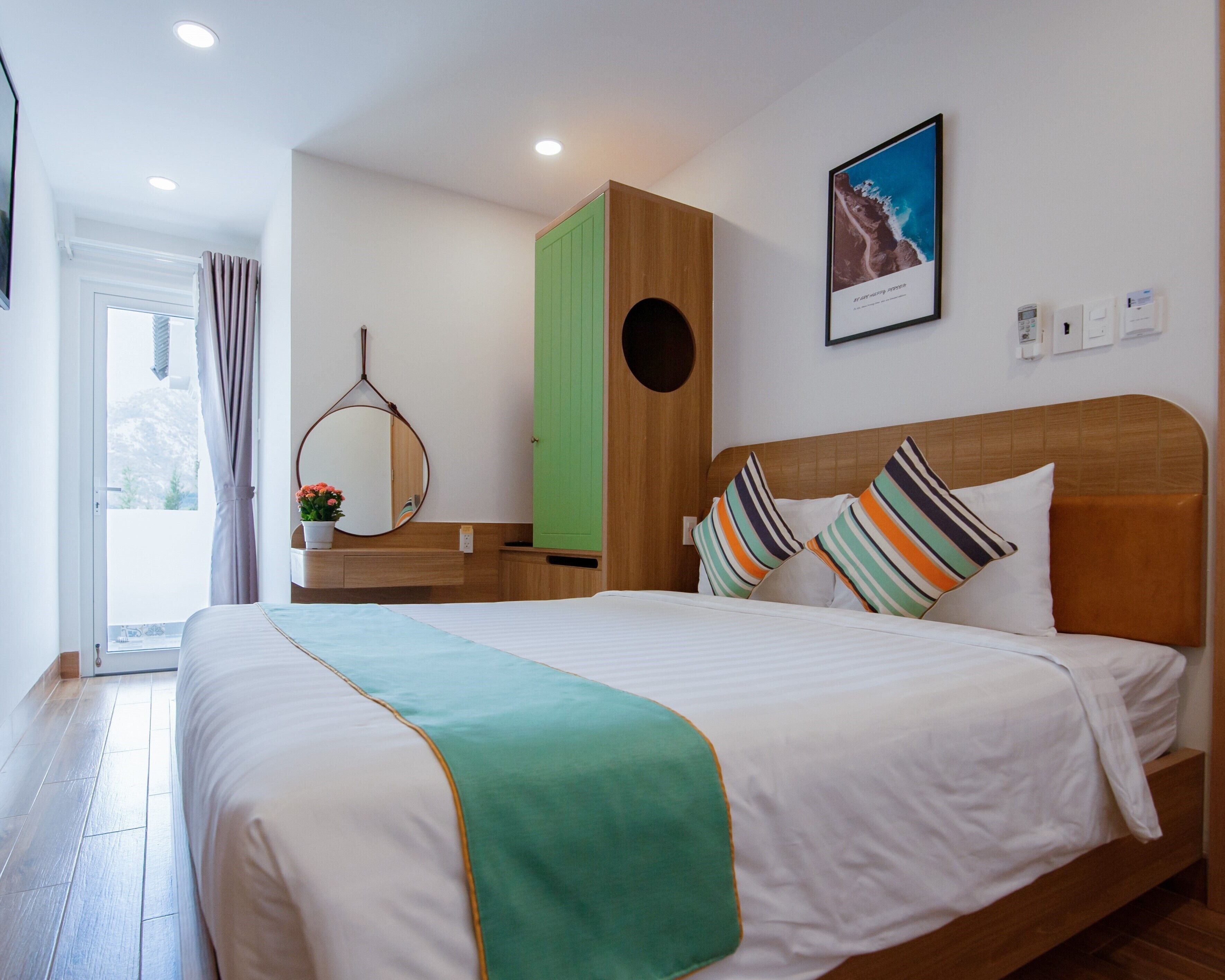 A neatly arranged hotel room with a bed, a green wardrobe, a mirror, and a framed picture on the wall.