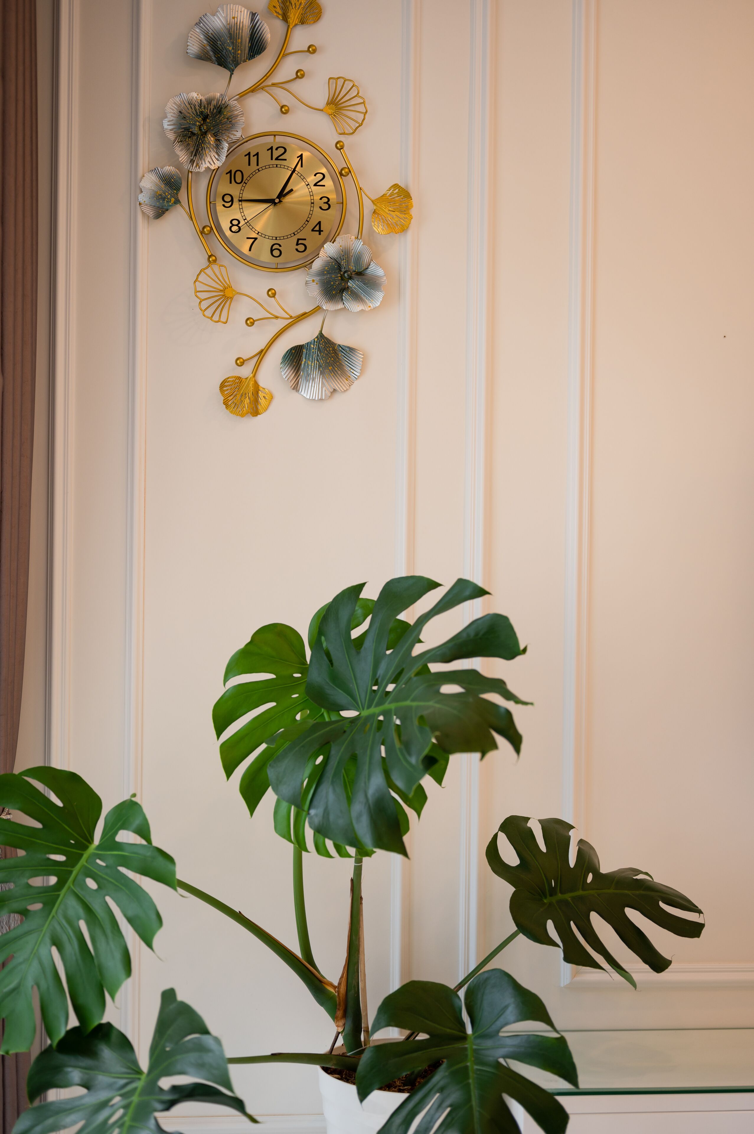 Un reloj montado en la pared con diseño floral, una planta de monstera en maceta y una pared blanca con paneles.
