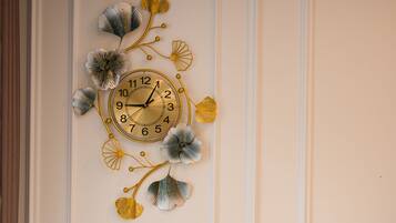 Un reloj montado en la pared con diseño floral, una planta de monstera en maceta y una pared blanca con paneles.