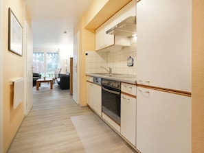 Un pasillo estrecho que conduce a una cocina con armarios blancos, un fregadero y un horno. Se ve un comedor al fondo con una mesa y sillas.