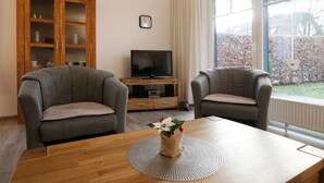 A living room with two grey armchairs, a wooden coffee table with a vase of flowers, a TV on a wooden stand, and a large window with a view of a garden. - Papillon Whg. 17/1, Germany (Ostseebad Boltenhagen)