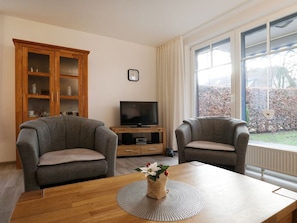 Una sala de estar con dos sillones grises, una mesa de centro de madera con un jarrón de flores, un televisor sobre un mueble de madera y un ventanal con vistas a un jardín.
