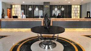 Een hotel receptie met een marmeren balie, een ronde tafel in het midden en een receptioniste achter de balie.