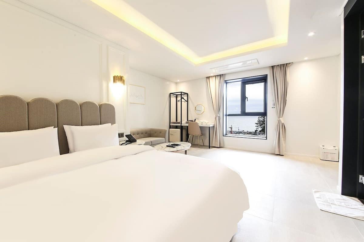 A modern hotel room with a large bed, a desk, and a chair. There is a window with curtains, a bedside table, and a lamp.