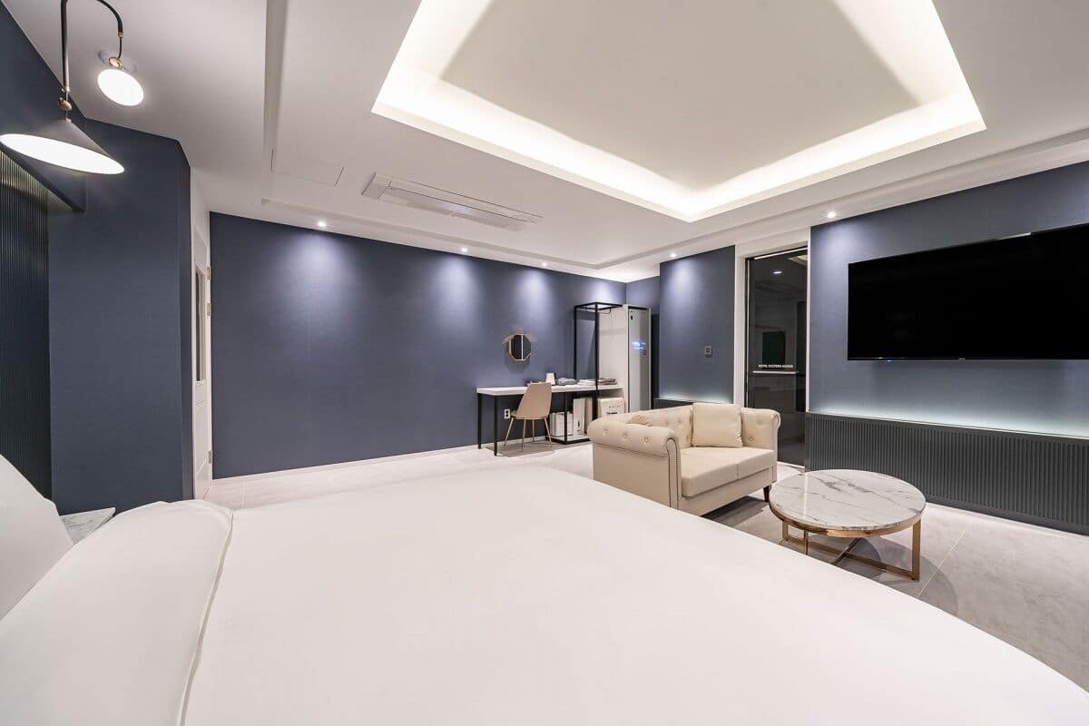 A modern hotel room with a large bed, a sofa, a round coffee table, a desk with a chair, a TV mounted on the wall, and a skylight.
