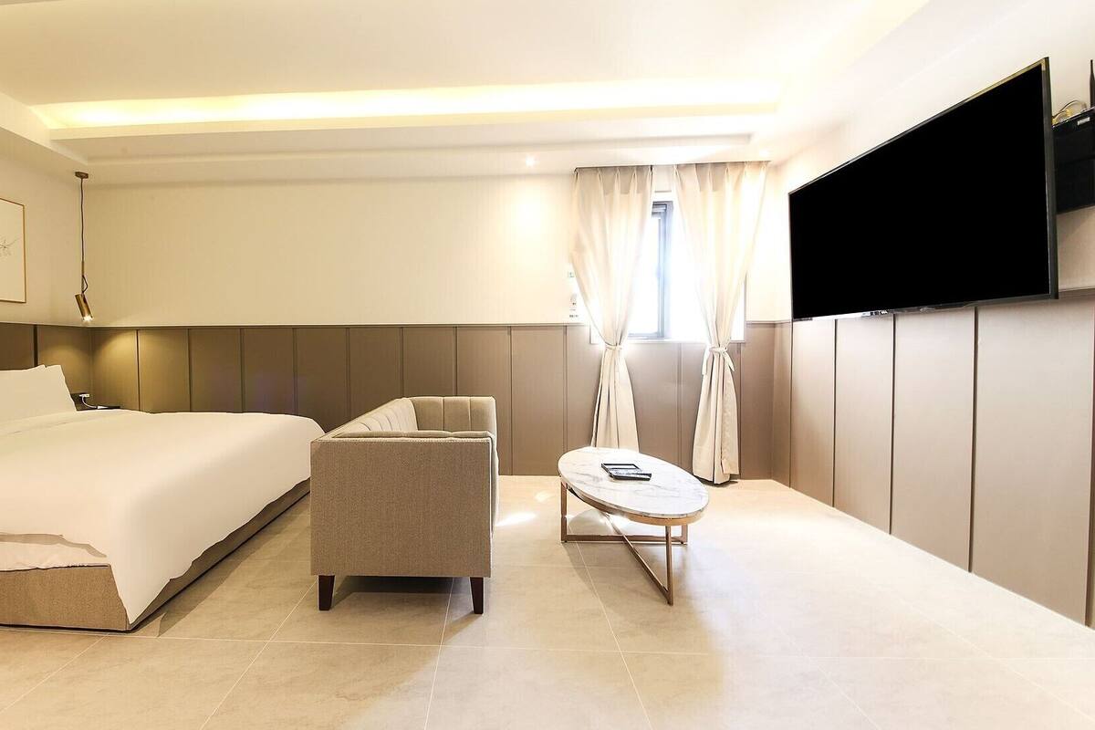 A modern hotel room with a large bed, a sofa, a small table, and a TV mounted on the wall.