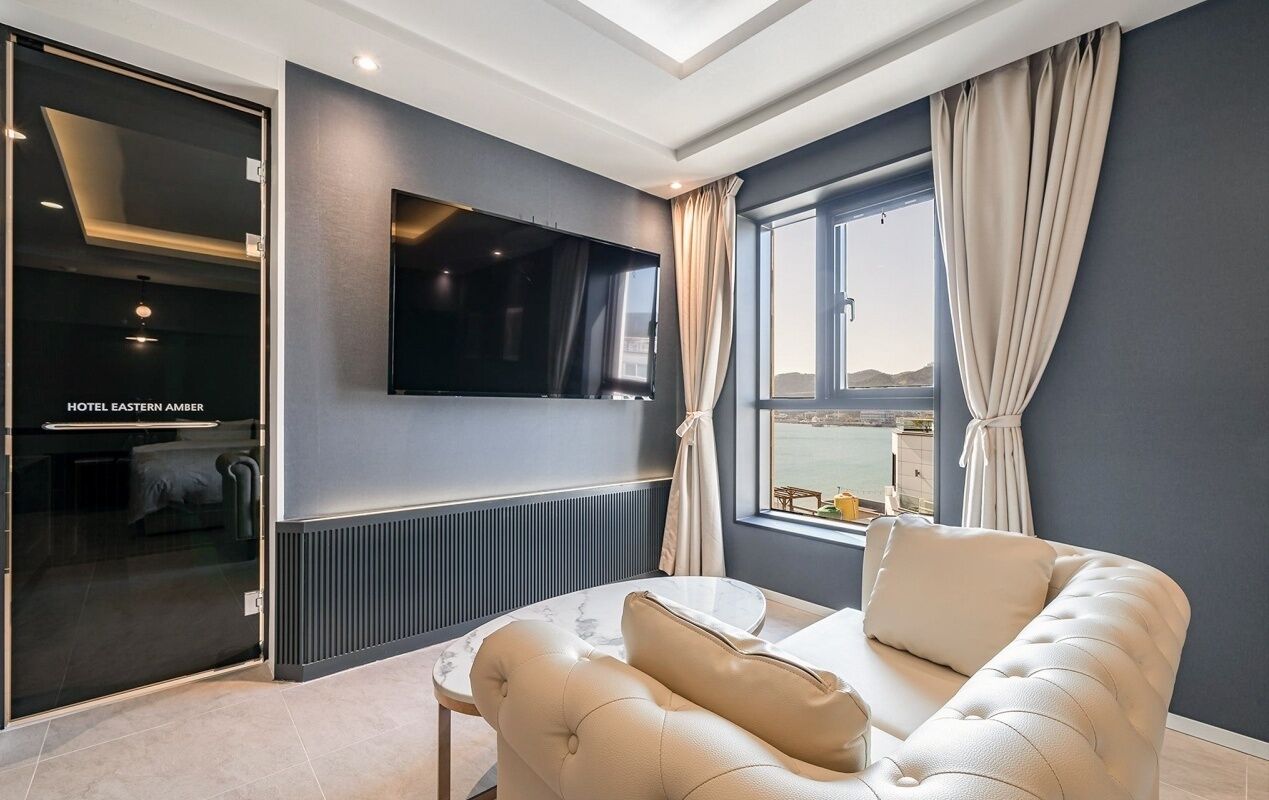 Ein modernes Hotelzimmer mit einem großen Flachbildfernseher an der Wand, einer gemütlichen Couch, einem Esstisch und Meerblick durch ein großes Fenster.