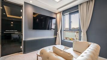 Ein modernes Hotelzimmer mit einem großen Flachbildfernseher an der Wand, einer gemütlichen Couch, einem Esstisch und Meerblick durch ein großes Fenster.
