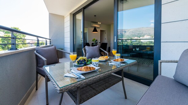 A balcony with a glass table set for breakfast, including croissants, fruit, and juice. - Apartment Luni (Dubrovnik)