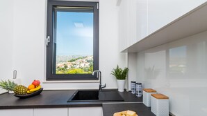 A modern kitchen with a black sink, a window with a view of a town, and a fruit bowl on the counter. - Apartment Luni (Dubrovnik)