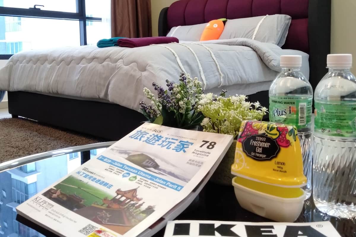 A hotel room with a bed, a window with curtains, a painting on the wall, and a table with magazines, a bottle of gel, and a water bottle.