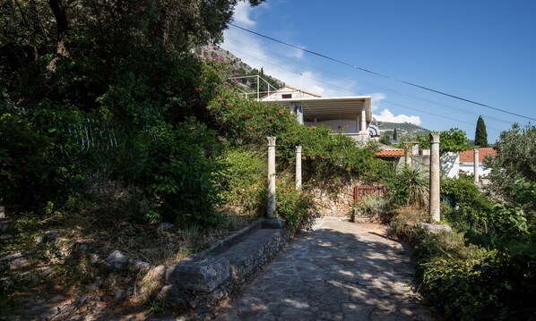 A stone pathway leading to a house with a red-tiled roof, surrounded by lush greenery and a clear blue sky.